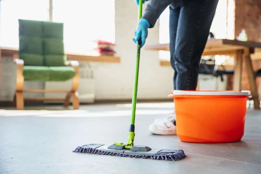 Person mopping a floor with a blue mop and orange bucket in a bright, modern workspace.